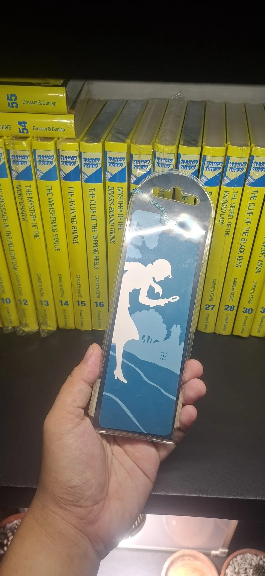 A silhouette-style Nancy Drew bookmark, a girl with a magnifying glass, held up beside the shelf
